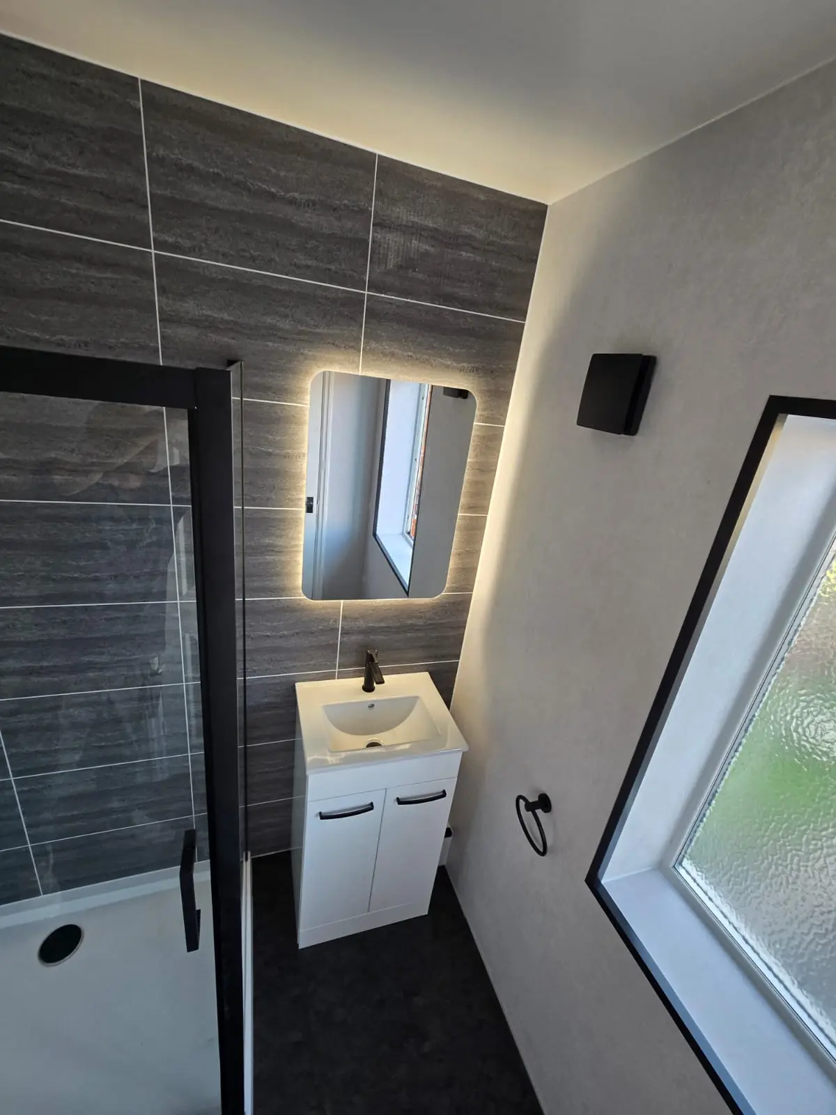 Backlit LED mirror above a white vanity with black tap on dark grey feature cladding in Sunderland